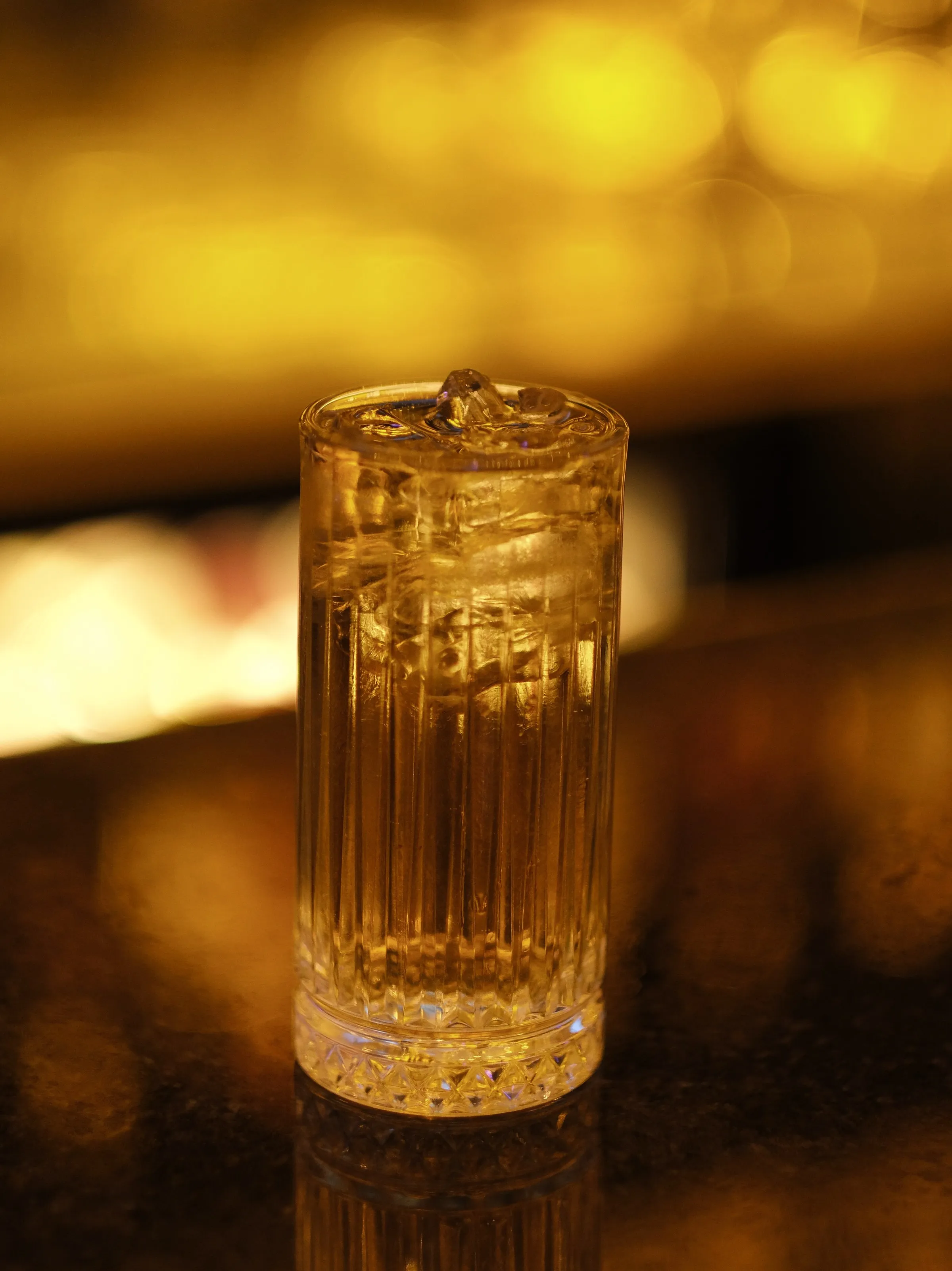 A whisky highball over ice in a tall ribbed glass, glowing gold in front of a warmly lit bar.