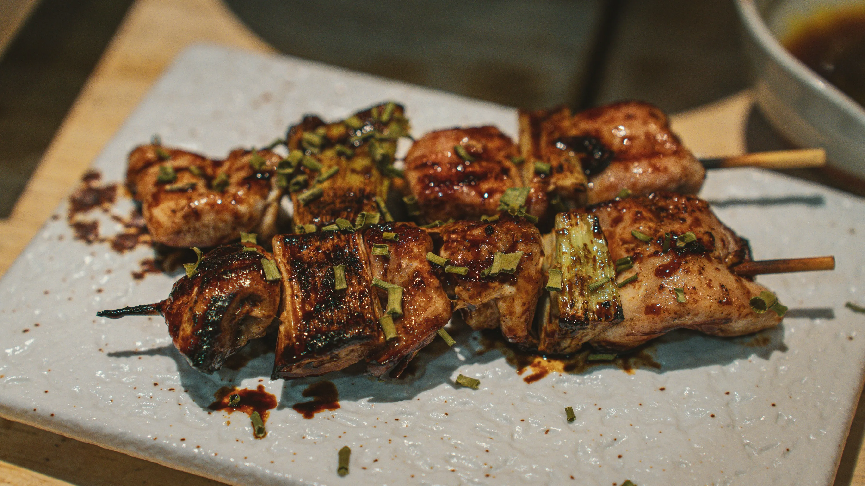 Two chicken and leek skewers, glazed with tare and scattered with chives, on a pale grey stone plate.